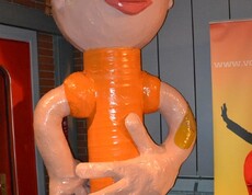 Eine Figur, die einem Menschen ähnelt, steht in einem Raum, ist überdimensioniert und orange und hautfarben. Sie hat überdies eine unproportionale linke Hand. 
