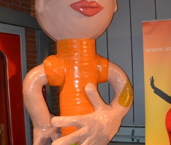 Eine Figur, die einem Menschen ähnelt, steht in einem Raum, ist überdimensioniert und orange und hautfarben. Sie hat überdies eine unproportionale linke Hand. 
