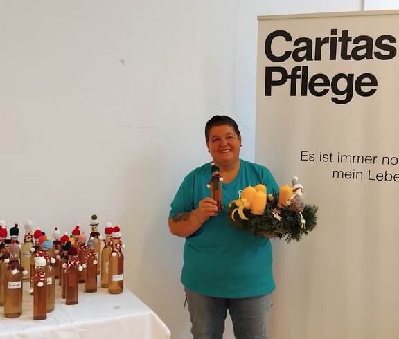 Eine Person steht neben einem Tisch mit dekorierten Flaschen und hält eine Flasche und einen Kranz mit Kerzen.