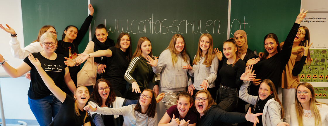 Auf der Tafel, vor der sich viele Schüler*innen der Caritas HLW in Klagenfurt versammelt haben, sich freuen und die Hände in die Luft strecken, steht die Website www.caritas-schulen.at.