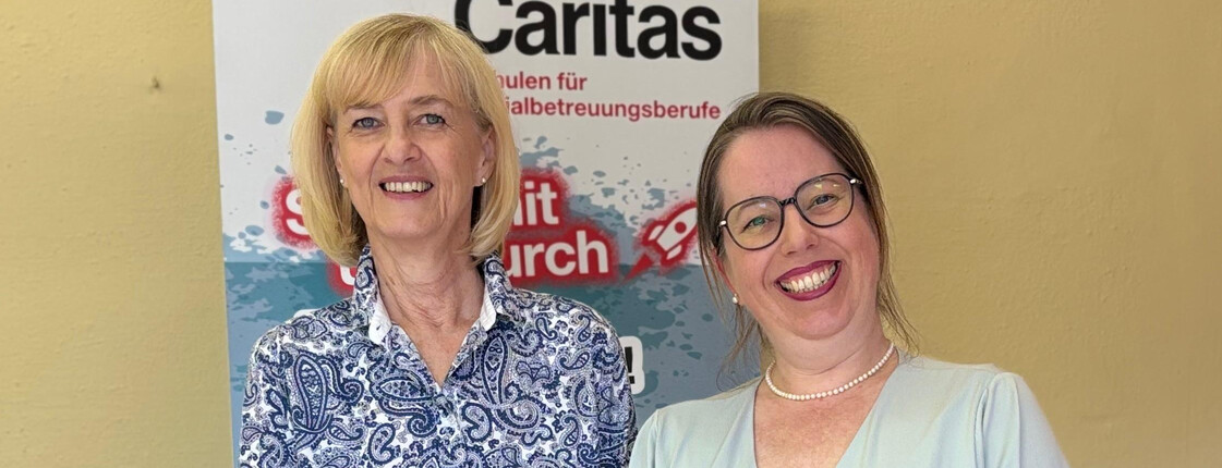 Zwei Frauen stehen vor einem Caritas Roll-Up und lächeln.