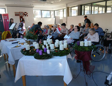 Mehrere Adventkränze mit Kerzen auf Tisch, Menschen sitzen dahinter und lesen