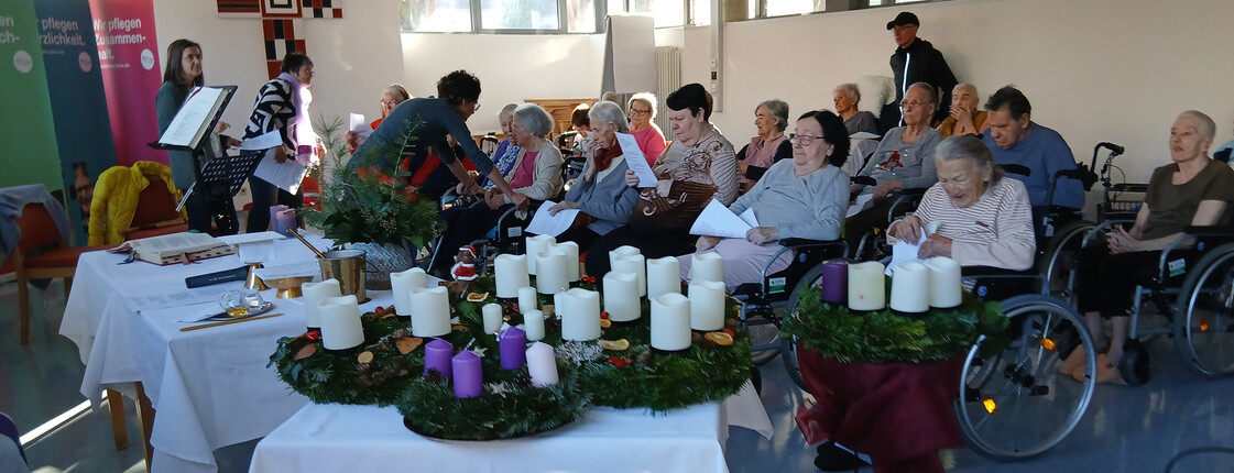 Mehrere Adventkränze mit Kerzen auf Tisch, Menschen sitzen dahinter und lesen