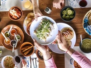 Vogelperspektive eines voll gedeckten Essenstisch aus Holz, an dem Personen sitzen und sich gegenseitig Schüsseln mit Essen reichen.