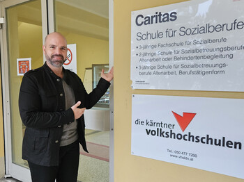 Schulleiter steht vor Eingang der Caritas-Schule für Sozialberufe in Wolfsberg und zeigt auf das Schulschild.