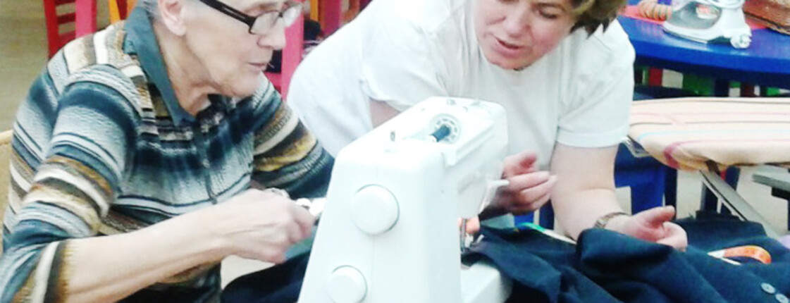 Zwei Damen sitzen an einem roten Tisch und reparieren mit einer Nähmaschine ein Kleidungsstück. Eine der beiden Frauen trägt ihre Haare kurz. Die andere hat lange graue Haare. 