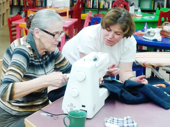 Zwei Damen sitzen an einem roten Tisch und reparieren mit einer Nähmaschine ein Kleidungsstück. Eine der beiden Frauen trägt ihre Haare kurz. Die andere hat lange graue Haare. 