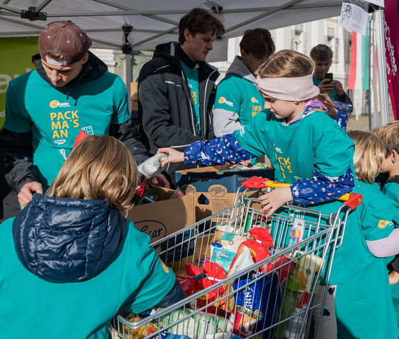 Kinder legen gespendete Lebensmittel in Kartons, unterstützt von Helfern der youngCaritas.