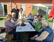 Mehrere Personen sitzen gemeinsam an einem Tisch auf der Terrasse und schneiden Maroni ein, während freiwillige Helferinnen und Helfer die Vorbereitungen für das anschließende Maronibraten unterstützen.