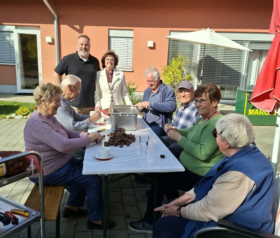 Mehrere Personen sitzen gemeinsam an einem Tisch auf der Terrasse und schneiden Maroni ein, während freiwillige Helferinnen und Helfer die Vorbereitungen für das anschließende Maronibraten unterstützen.
