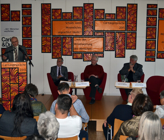 Bei den 13. Caritasgesprächen disskutierten die Referenten mit Einbindung des Publikums das Thema Schuld. Das Bild zeigt das von hinten fotografierte Publikum und die auf der Bühne stehenden bzw. sitzenden Referenten. Geschmückt wurde der Saal mit oranger Farbe. 