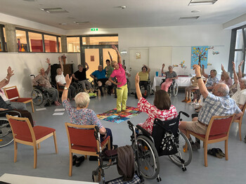 Senior*innen heben im Kreis sitzend die Arme bei einer Gruppenübung im Pflegewohnhaus „Haus Martha“.