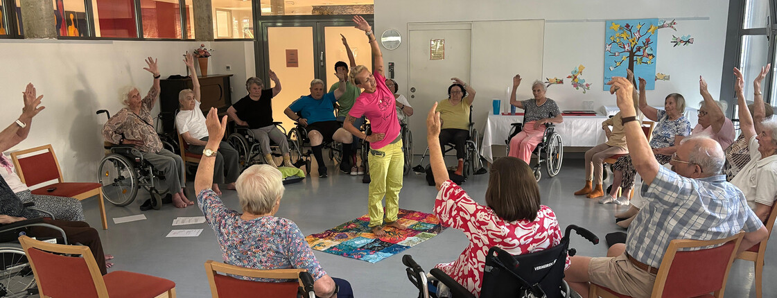 Senior*innen heben im Kreis sitzend die Arme bei einer Gruppenübung im Pflegewohnhaus „Haus Martha“.