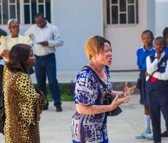 Eine Trainerin von FLEM spricht gestikulierend zu einer Gruppe; im Hintergrund stehen Erwachsene sowie Schülerinnen und Schüler in Schuluniform, die aufmerksam zuhören.