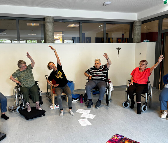 Gruppe älterer Menschen im Rollstuhl führt synchron eine Arm- und Rumpfdehnung durch.
