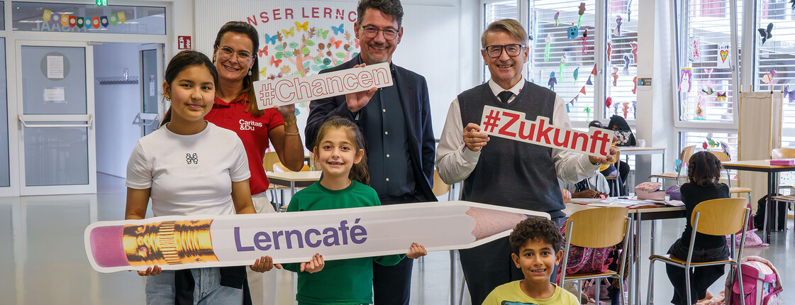 Zwei Männer, eine Caritas-Mitarbeiterin und drei Kinder halten Lerncafé-Schilder in einem Lernraum.