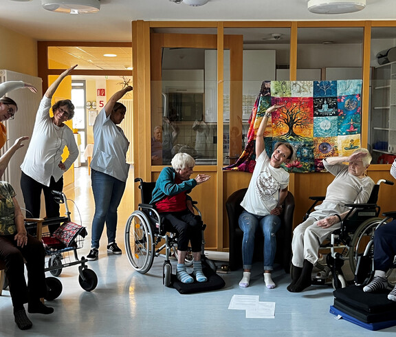 Pflegekraft leitet Senior*innen im Rollstuhl zu einer achtsamen Bewegungseinheit im Aufenthaltsraum an.