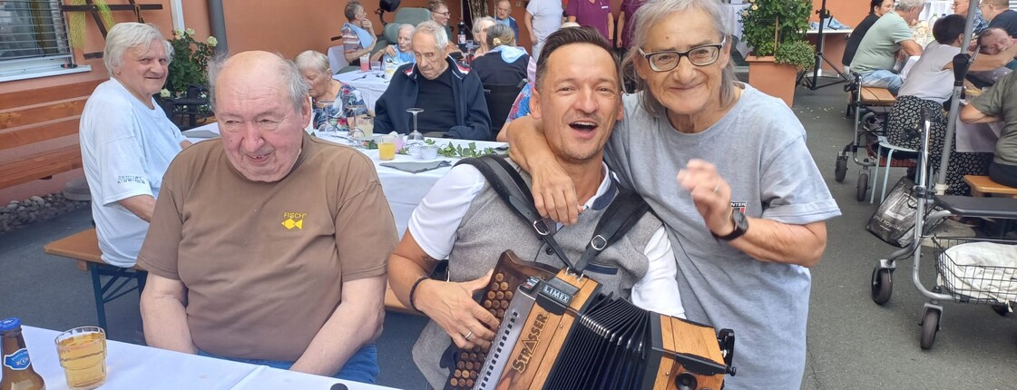 Eine Person mit einer Ziehharmonika sitzt zwischen den Gästen des Sommerfestes, während die Bewohnerinnen und Bewohner an festlich gedeckten Tischen im Innenhof zusammensitzen und der Musik lauschen.