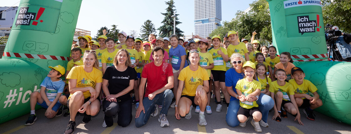 Viele Erwachsene und Kinder in Lauf-Gewand winken lächelnd unter der Startlinie der Laufstrecke eines Benifiz-Laufes.