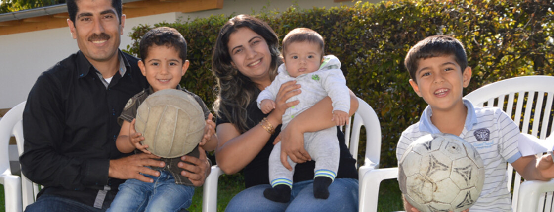 Eine junge Familie mit drei Kindern sitzt in einem Garten auf Plastikstühlen. Sie wirken glücklich und zwei der jungen Buben halten einen Fußball in der Hand. 