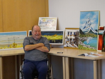 Bilder stehen im Hintergrund auf einem Tisch und lehnen an der Wand. Davor im Rollstuhl sitzt ein Mann mit den Armen verschränkt vor der Brust.