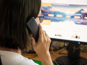 Eine Braunhaarige mit Brille hält einen Telefonhörer in der Hand und sieht dabei auf den Bildschirm, auf dem man die Website der Telefonseelsorge Österreich leicht verschwommen sieht.