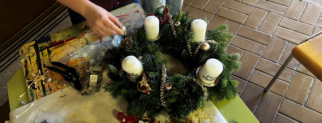 Ein Adventkranz wird auf einem Tisch geschmückt, rundherum liegen Schmuck und eine Heißklebepistole, man sieht die Hände von einer Schülerin, die am Adventkranz arbeitet.