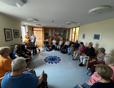 Senior*innen sitzen im Kreis um ein rundes Tuch mit Pfauenfeder-Muster und üben stille Meditation.