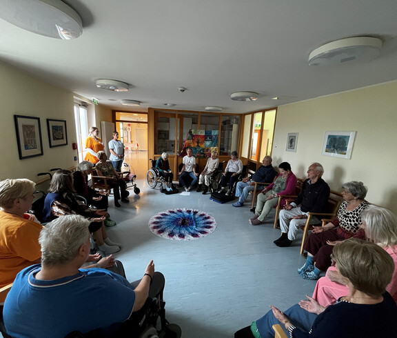 Senior*innen sitzen im Kreis um ein rundes Tuch mit Pfauenfeder-Muster und üben stille Meditation.