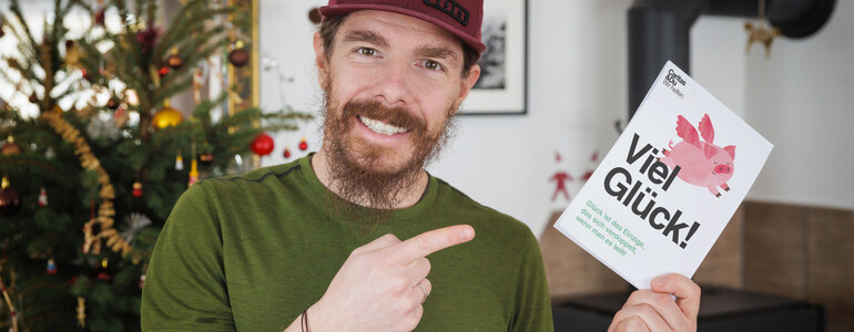 Ein Mann lächelt in die Kamera und hält eine Karte auf der "Viel Glück" steht.
