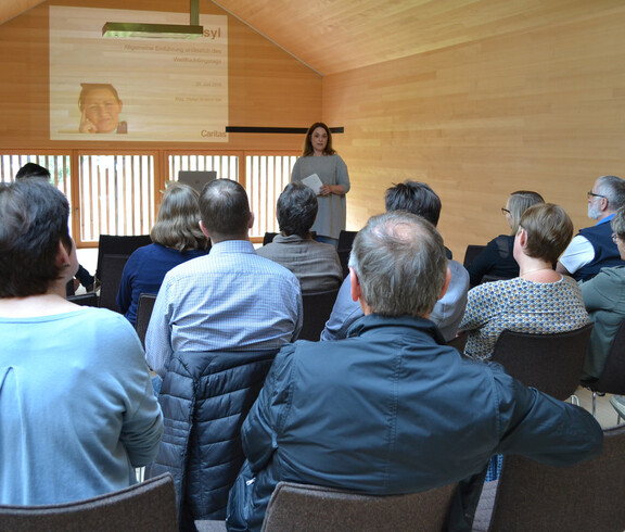 Eine junge Frau hält eine Präsentation in einem Raum aus Holz. Das Publikum sitzt auf Stühlen und blick in ihre Richtung.