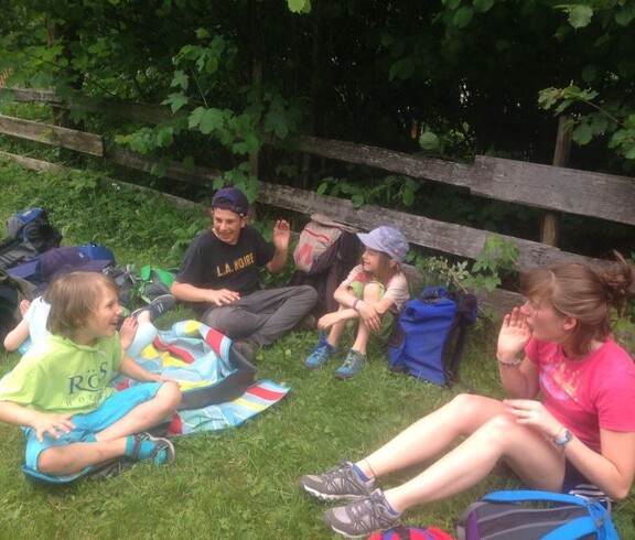 Kinder sitzen während einer Projektwoche auf einer Wiese am Waldrand, machen eine Pause und verbringen gemeinsam Zeit im Freien.