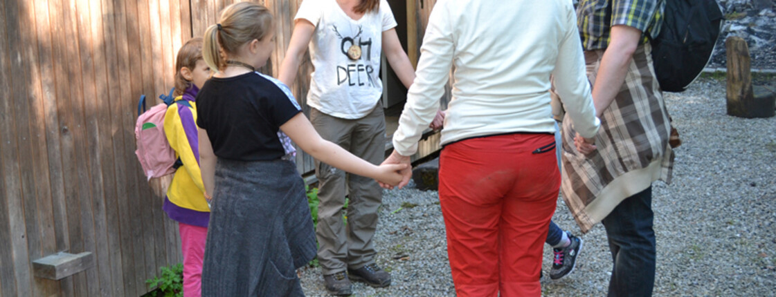 Drei Kinder stehen mit drei Erwachsenen in einem Kreis im Freien zusammen. Dabei halten sich alle am Arm.
