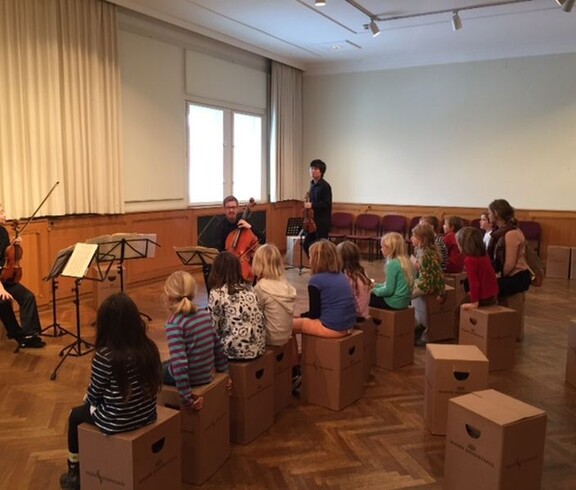 Kinder sitzen bei einer musikalischen Präsentation und erleben die Klänge von Geige und Cello in einem Veranstaltungsraum.