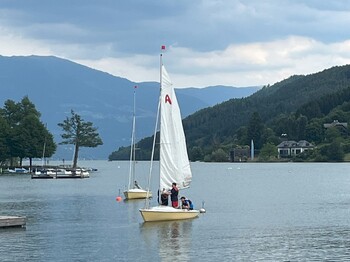 Kleine Segelboote liegen auf einem Bergsee vor bewaldeten Hängen. Die Szene zeigt Wassersport in einer ruhigen alpinen Landschaft.