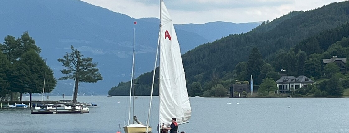 Kleine Segelboote liegen auf einem Bergsee vor bewaldeten Hängen. Die Szene zeigt Wassersport in einer ruhigen alpinen Landschaft.