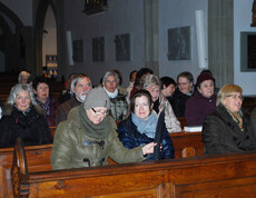 Etliche Damen mittleren Alters und vereinzelt auch Herren sitzen in Kirchenbänken. Sie sind alle warm bekleidet.  