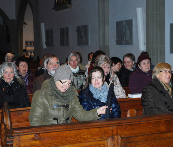 Etliche Damen mittleren Alters und vereinzelt auch Herren sitzen in Kirchenbänken. Sie sind alle warm bekleidet.  