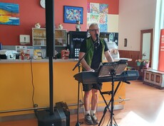 Eine Person steht an einem Keyboard mit Mikrofon und Lautsprecheranlage und sorgt in einem hellen Aufenthaltsraum mit farbenfrohen Wandbildern und Kunstwerken für die musikalische Unterhaltung der Veranstaltung.