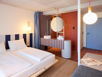 Innenansicht eines Hotelzimmers mit Doppelbett, integriertem offenem Waschbereich, rundem Spiegel, rot-blauen Fliesen, blauen Vorhängen und hellem Holzboden. Im Hintergrund befinden sich Eingangsbereich und Zimmertür.