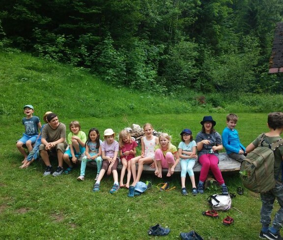 Kinder sitzen während der Projektwoche gemeinsam auf einer Holzbank im Freien und verbringen eine Pause in naturnaher Umgebung.