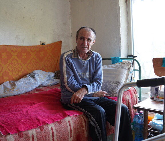 Ein älterer Mann sitzt auf einem Sofabett und blickt in die Kamera.