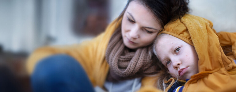 Eine Mutter sitzt mit verzweifeltem Gesichtsausdruck mit ihrem in eine Decke eingewickelten Kind auf einer Couch.