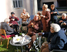 Ältere Menschen, einige in Rollstühlen, genießen eine Pause bei Tee oder Kaffee im Garten.