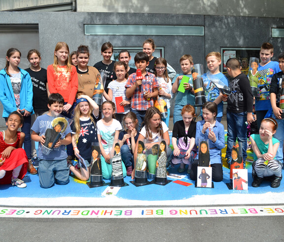 Kinder unterschiedlichen Alters stehen auf einer großen Landkarte, die den ganzen Globus repräsentiert. In der Mitte steht ein Bub, der ein ORF Mikrofon in der Hand hält. 