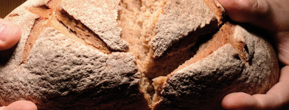 Zwei Hände halten ein großes Brot in der Hand. Es erleuchtet auf schwarzem Hintergrund.