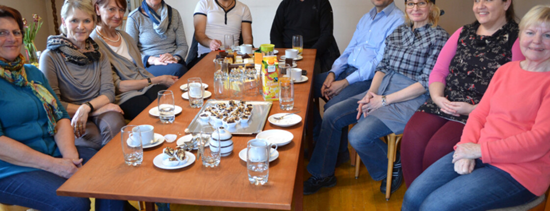 Mehrere Menschen sitzen in einer Runde um eine lange Tafel. Auf dem Tisch befinden sich Snacks und Getränke.
