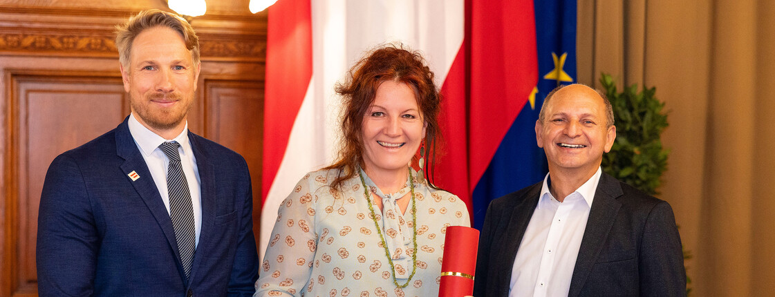 Eine Caritas Mitarbeiterin steht zwischen zwei Männern in Anzügen. Im Hintergrund sieht man die Flagge Österreichs und der Europäischen Union.