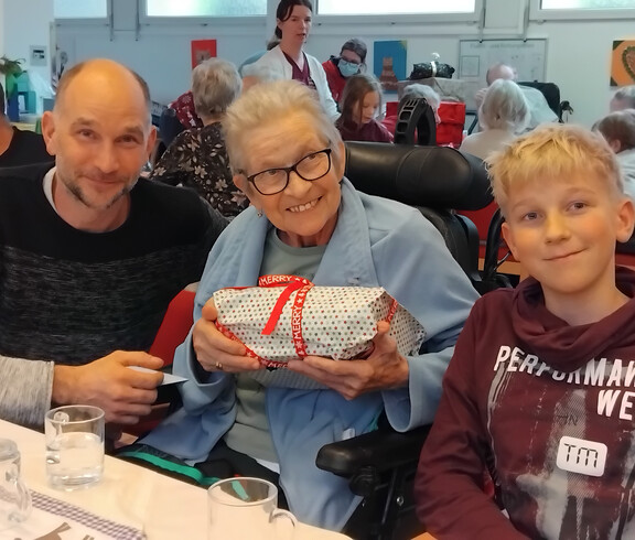 GeschenkBewohner*in im Rollstuhl hält ein verpacktes Geschenk, flankiert von zwei Angehörigen